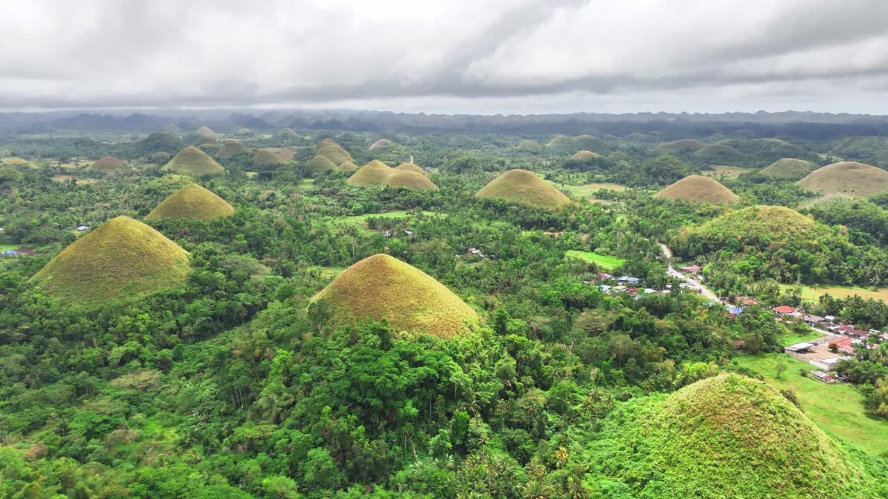 Drone reveals the famed Chocolate Hills in Bohol, Philippines—symmetrical grass-covered mounds spread across a lush tropical landscape, with distant villages and cloudy skies