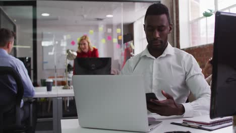 African-american-businessman-sitting-at-desk-talking-on-smartphone-using-laptop