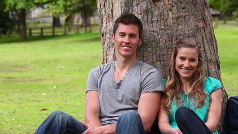 camera pans as two friends sit against a tree while smiling and looking at the camera