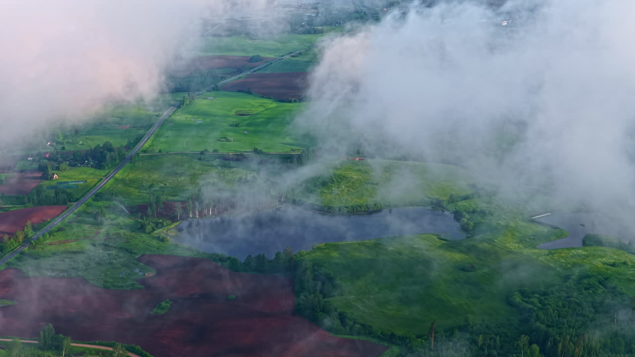 A breathtaking aerial drone shot passes through low-hanging clouds to reveal a stunning view of a tranquil lake surrounded by a patchwork of green and brown fields in the rural Latvian countryside