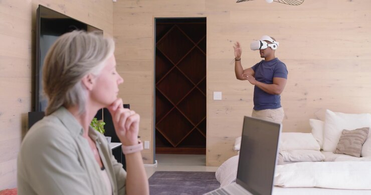 Coppia multietnica a casa, l'uomo indossa un visore VR bianco gesticolando per interagire, la donna lavora al laptop.