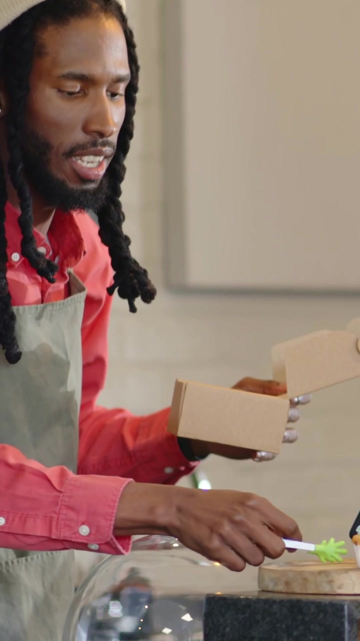 Vertical video: Packing barista prompted by order placing cupcake into box at cafe in gray apron