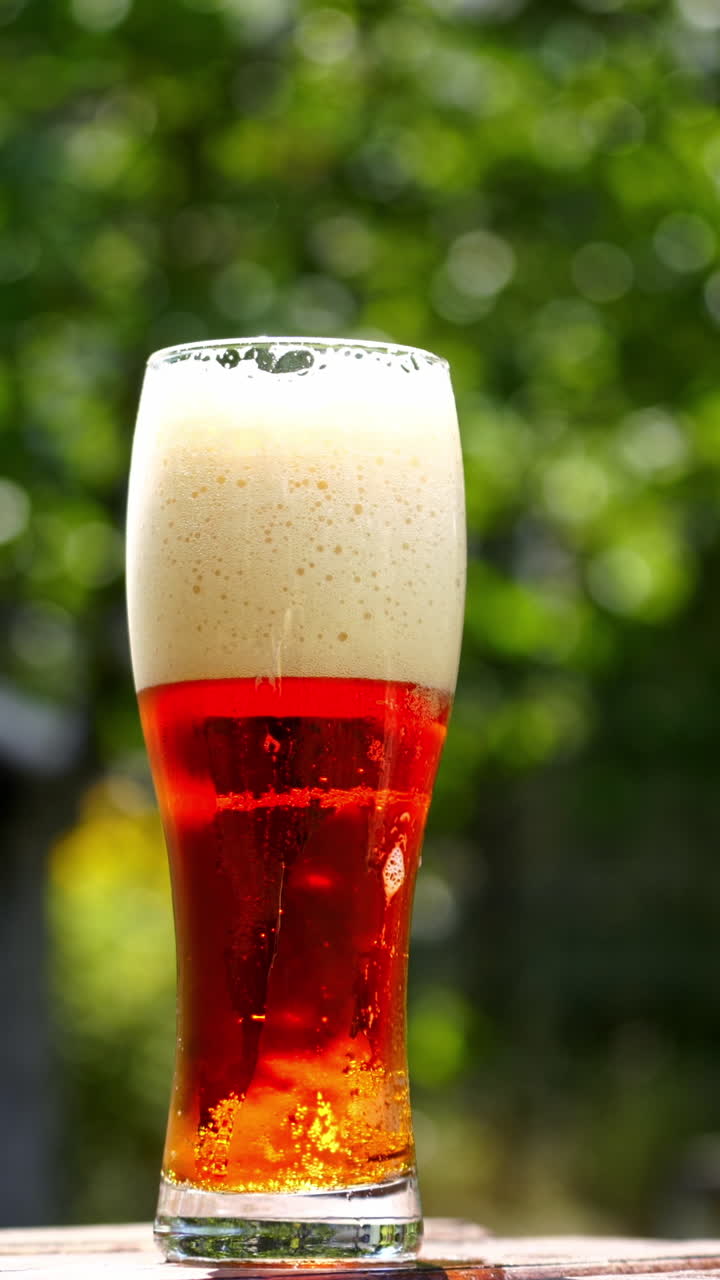 Cold beer in a glass with white foam. Craft beer in a glass with small bubbles inside. Cool brown beverage on a table outdoors. Close-up. Vertical video