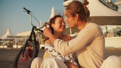 Happy-couple-on-a-date,-a-blond-guy-in-light-clothes-straightens-the-hair-of-a-brown-haired-girl-in-light-clothes-with-a-black-bow-who-sits-near-a-bicycle-on-the-beach