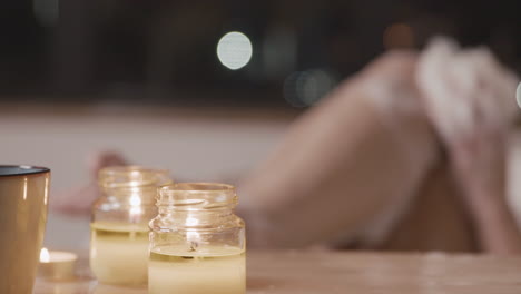 vista de cerca de una mesa con velas e incienso, en el fondo una mujer borrosa tomando un baño mientras se frota las piernas con jabón