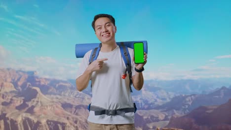 asian male hiker with mountaineering backpack smiling and pointing to mock up green screen smartphone while traveling at the top of mountain