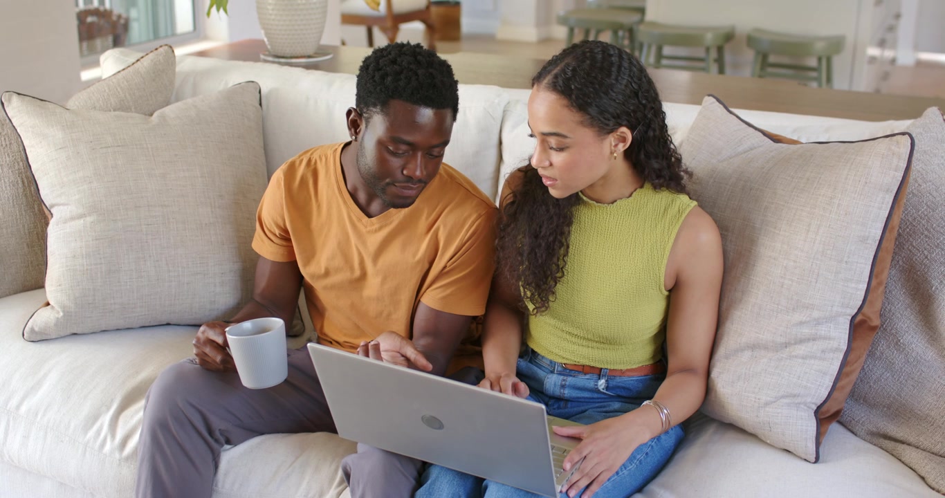 Vertical video: African American couple on sofa, man with mug tapping laptop, woman navigating web