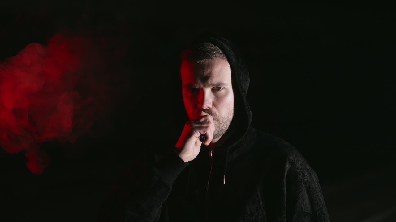 a man smokes an e cigarette. he wears a hooded sweatshirt. in the background fog. his face is half illuminated in red with a dark background