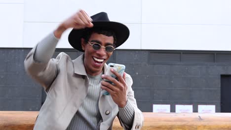 Happy-stylish-ethnic-man-watching-mobile-phone-while-sitting-on-bench