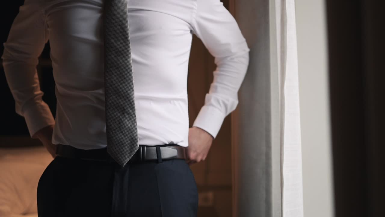 Uomo che si sistema la cravatta e la cintura, vestito in abito formale con una camicia bianca e una cravatta grigia.