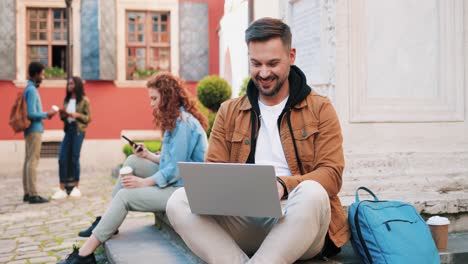 Caucasian-bearded-man-using-laptop-while-sitting-on-the-ground-in-the-street