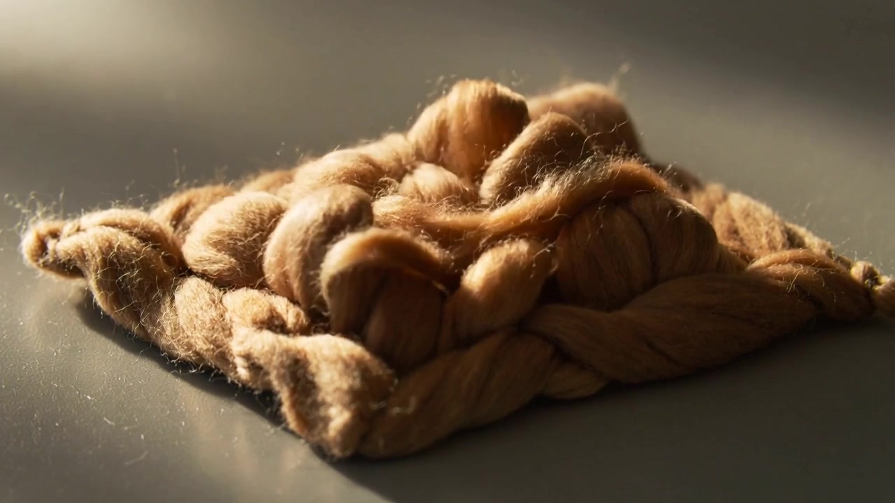 Beiges geflochtenes Polster reagiert auf kurze Krafteinwirkung, hebt eine geschlungene Quaste auf grauer Tischplatte an, Staubpartikel.