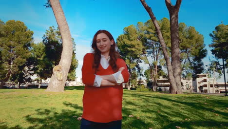 una feliz, atractiva, joven, mujer caucásica en el parque, se vuelve hacia la cámara, cruza los brazos, y sonríe con gran confianza