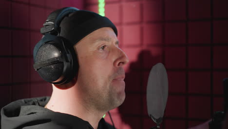 close-up side shot of a man in black wearing headphones, standing in front of a microphone in a soundproofed room. he is singing with seriousness, accompanied by expressive hand movements