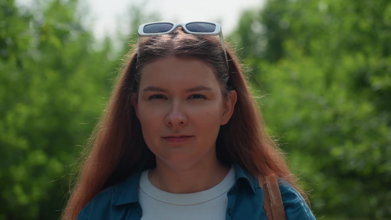 Donna in abito bianco che si mette gli occhiali da sole in testa mentre si trova in un viale verde del parco, indossa una camicia blu e un abito bianco, circondata da un fogliame estivo luminoso, viso sereno, movimento rilassato, calda luce solare