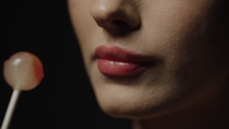 vista de cerca de una seductora rubia joven mujer caucásica con labios rojos lamiendo una deliciosa piruleta dulce y disfrutando del caramelo contra un fondo negro en cámara lenta