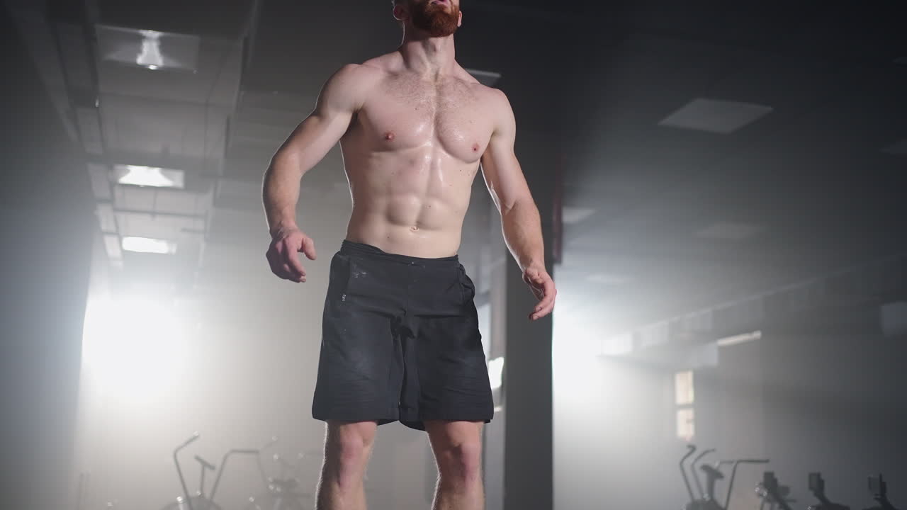 슬로우 모션: 근육이 있는 셔츠 없는 체력 있는 남자가 하드코어 체육관에서 힘차게 박스 점프를 하며 크로스 피트니스 훈련 계획의 일부를 하고 있습니다. 남자는 격렬한 운동으로  ⁇ 을 흘리고 있습니다.