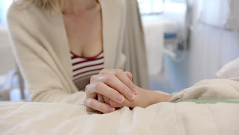 Midsection-of-caucasian-mother-holding-hand-of-daughter-patient-lying-in-hospital-bed,-slow-motion