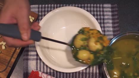 cook serving a veggie soup onto a white plate on the kitchen table