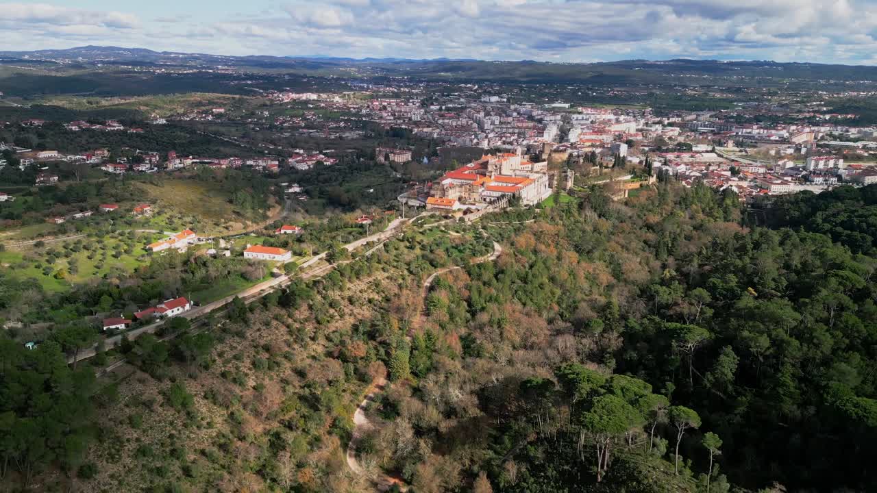 Ampla e épica vista de drone descendente do Convento de Cristo, acima de Tomar, Portugal, que combina a arquitetura histórica com a mata verdejante e a vasta paisagem urbana ao fundo.