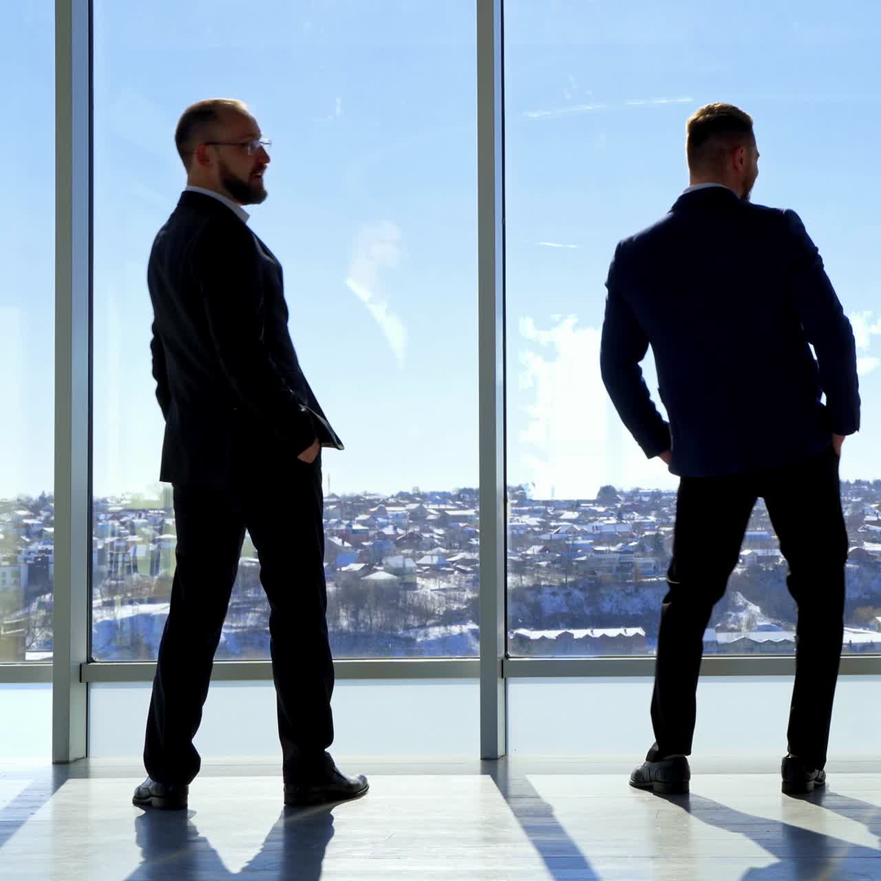Empresarios divertidos cerca de las ventanas de la oficina. Hombres con trajes elegantes divirtiéndose mientras miran a través de grandes ventanas. Vista trasera.