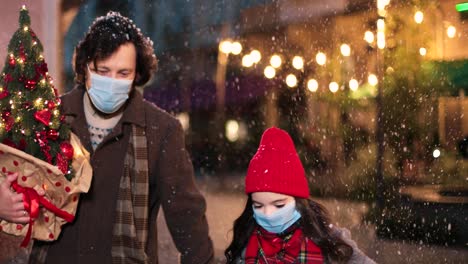 close-up view of caucasian happy father and daughter wearing facial masks walking down the street and holding christmas tree and present while it¬¥s snowing in christmas