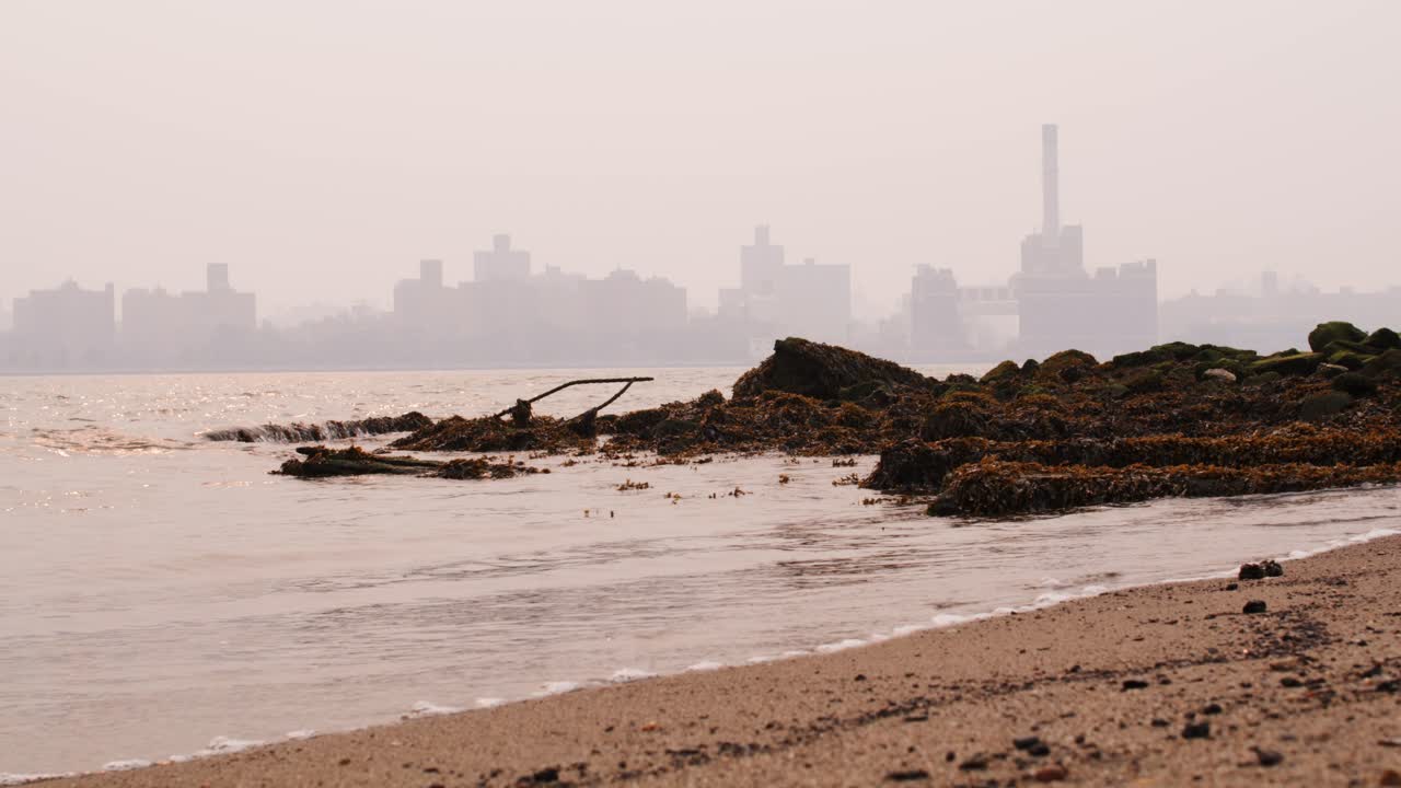 View of Manhattan covered in smoke from wildfires seen from beach on the east river with waves crashing on sandy beach and mossy rocks in the foreground