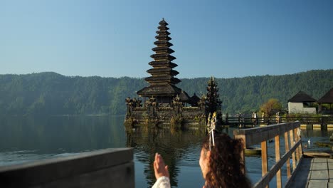 Ascending-slow-motion-shot-of-a-young-traveler-sitting-on-the-shore-danau-batur-in-front-of-the-water-temple-Pura-Ulun-Danu-temple-overlooking-the-beautiful-lake-with-beautiful-nature