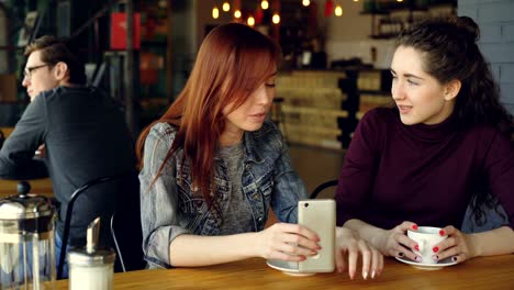 two attractive women in casual clothes are having girl time in cafe talking and holding coffee cup and smartphone. nice cafe interior, customers and furniture are visible.