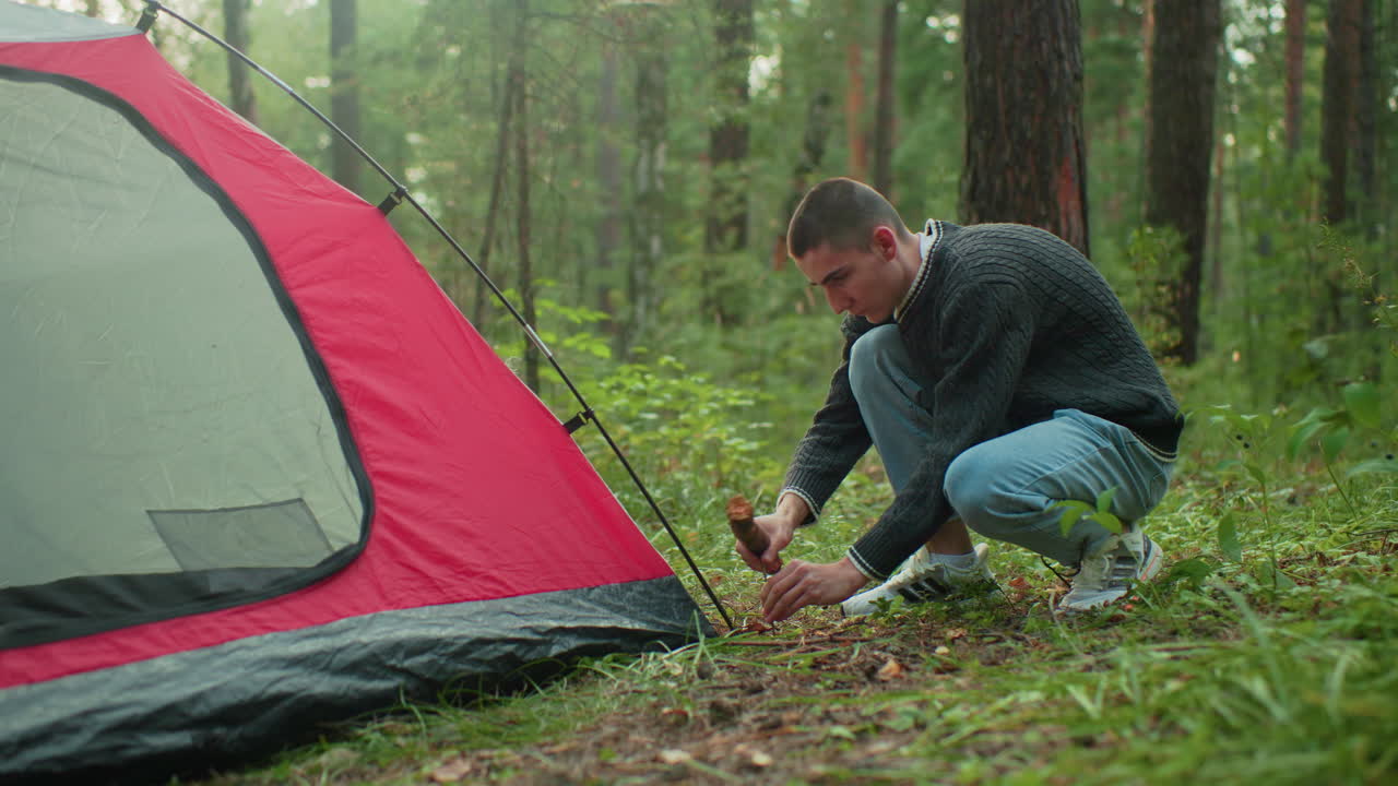 camper squats beside tent holding thick stick while hammering peg into grassy forest ground focused on securing tent with sunlight filtering through trees during peaceful outdoor setup