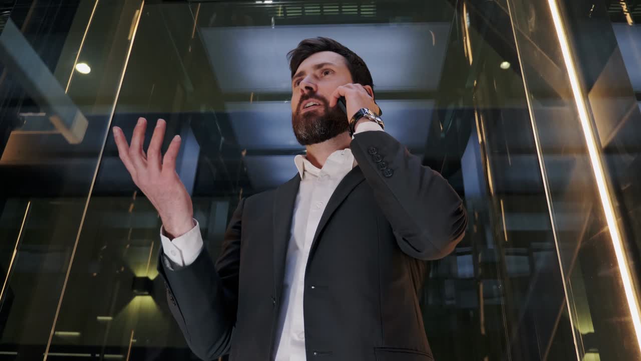 харизматичный успешный бизнесмен в костюме едет на стеклянном лифте в современном бизнес-центре. красивый серьезный мужчина разговаривает по мобильному телефону с деловым звонком в лифте. мужской лидер использует смартфон.