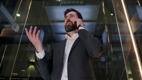 hombre de negocios carismático exitoso en un traje montando un ascensor de vidrio en un centro de negocios moderno. hombre guapo y serio hablando por teléfono móvil teniendo una llamada de negocios en un ascensor. líder masculino usando teléfono inteligente.