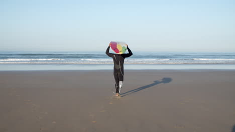 back view of a male surfer with prosthetic leg walking to the ocean with surfboard on his head