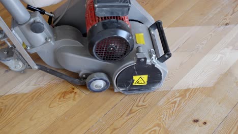 powerful industrial floor sander moves across aged pine floorboards, removing old finish and smoothing imperfections, with the machine’s red motor and yellow cord prominent, real time static camera