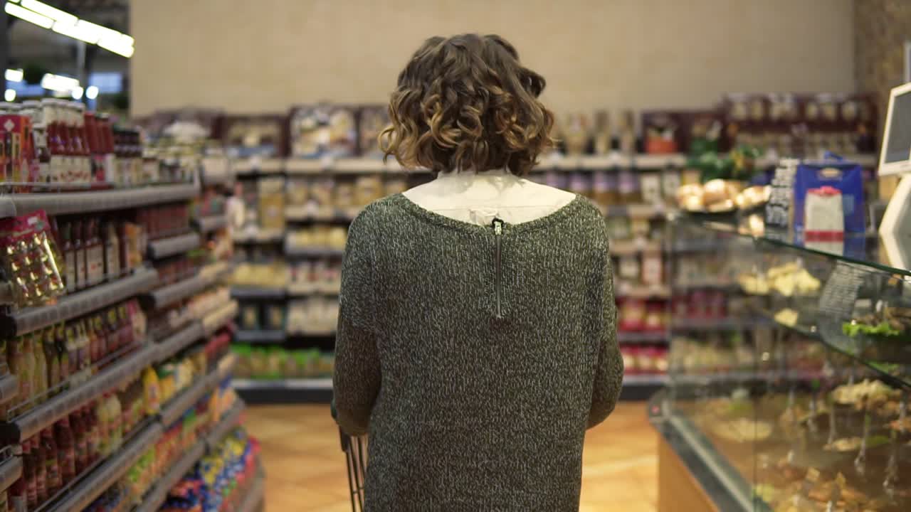 vista rara de una mujer con cabello ondulado está conduciendo el carrito de compras a través del departamento de alimentos en el supermercado y mirando a su alrededor. chicas bonitas, la vida cotidiana y la compra de productos concepto