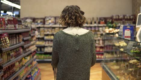 Rare-view-of-a-woman-with-wavy-hair-is-driving-shopping-trolley-through-food-department-in-supermarket-and-looking-around.-Pretty-girls,-everyday-life-and-buying-products-concept