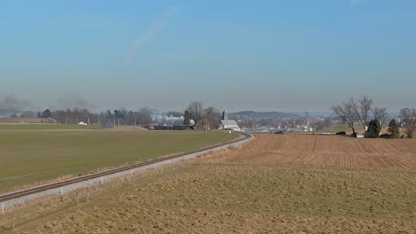 Una-Vista-Aérea-De-Un-Tren-De-Pasajeros-De-Vapor-Que-Se-Acerca,-Soplando-Humo,-Mientras-Viaja-Por-El-Campo,-En-Un-Soleado-Día-De-Invierno,-En-Un-Soleado-Día-De-Invierno