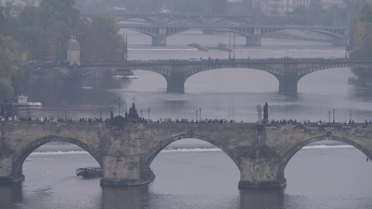 Wide shot of Prague's bridges on a foggy day, with people walking on the bridge and boats sailing on the water