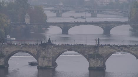 Wide-shot-of-Prague's-bridges-on-a-foggy-day,-with-people-walking-on-the-bridge-and-boats-sailing-on-the-water