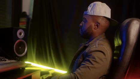 handsome young man sound producer in a hat sitting in the music studio at the mixer and working, then turning head to the camera and smiling cheerfully