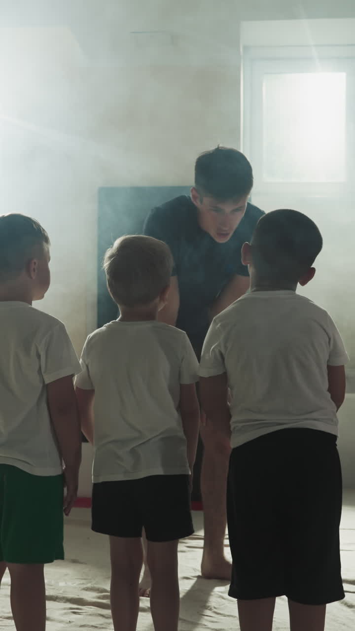 무술 체육 수업에서 젊은 트레이너와 어린이 그룹, 호기심 많은 어린 아이들이 안개 체육관에서 느린 동작으로 운동하기 전에 센세이를 봅니다.