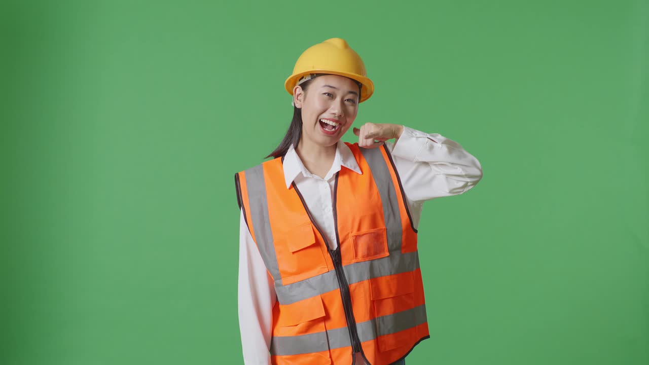 Asian Female Engineer With Safety Helmet Smiling To Camera And Making Call Me Gesture While Standing In The Green Screen Background Studio