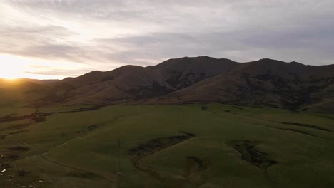 aerial - green meadows at the foot of rugged, brown mountains at sunrise