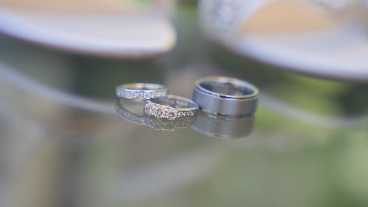 Eine detaillierte Makroaufnahme, die einen funkelnden Diamant-Verlobungsring, einen passenden Ehering und einen Herrenring aus gebürstetem Metall zeigt, die auf einem Glastisch mit sanftem Bokeh liegen
