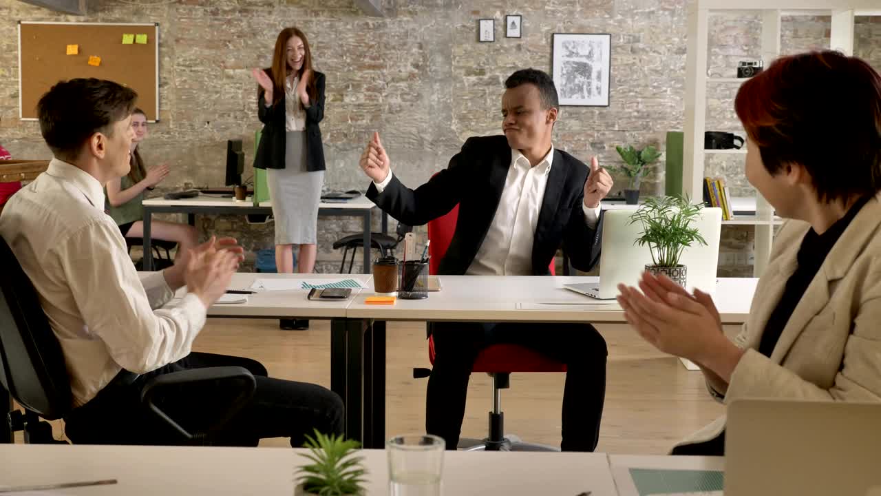 사무실에서 일하는 혼합된 민족의 사업가들, 그리고 갑자기 흑인 남자가 환호하고 춤추기 시작하고, 모두가 박수를 치고 흥분합니다.