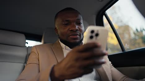 confident man businessman with black skin color with a light brown beard in messenger on his white phone while driving in a modern interior in a car around the city in summer