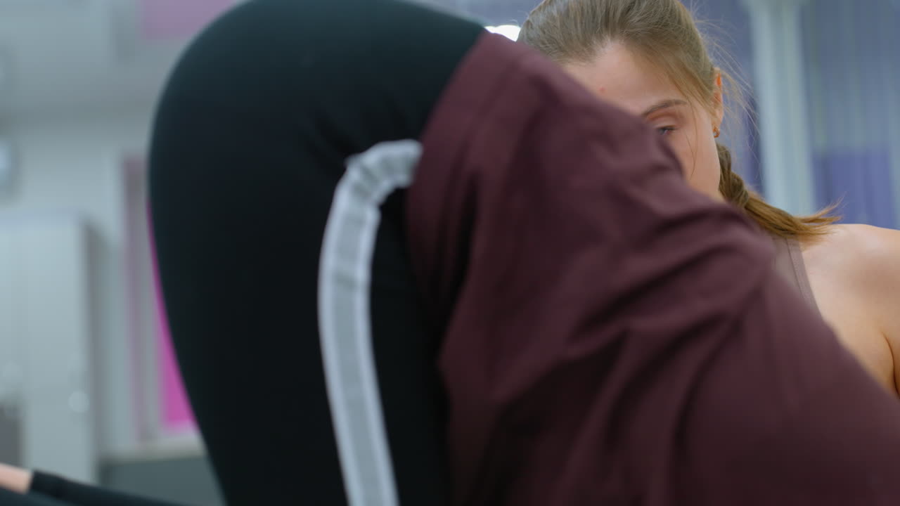Primo piano di una persona che fatica a bilanciarsi su una palla da ginnastica verde mentre è assistita da un istruttore, lo sfondo presenta decorazioni di armadietti e un grande specchio che riflette l'ambiente della palestra.
