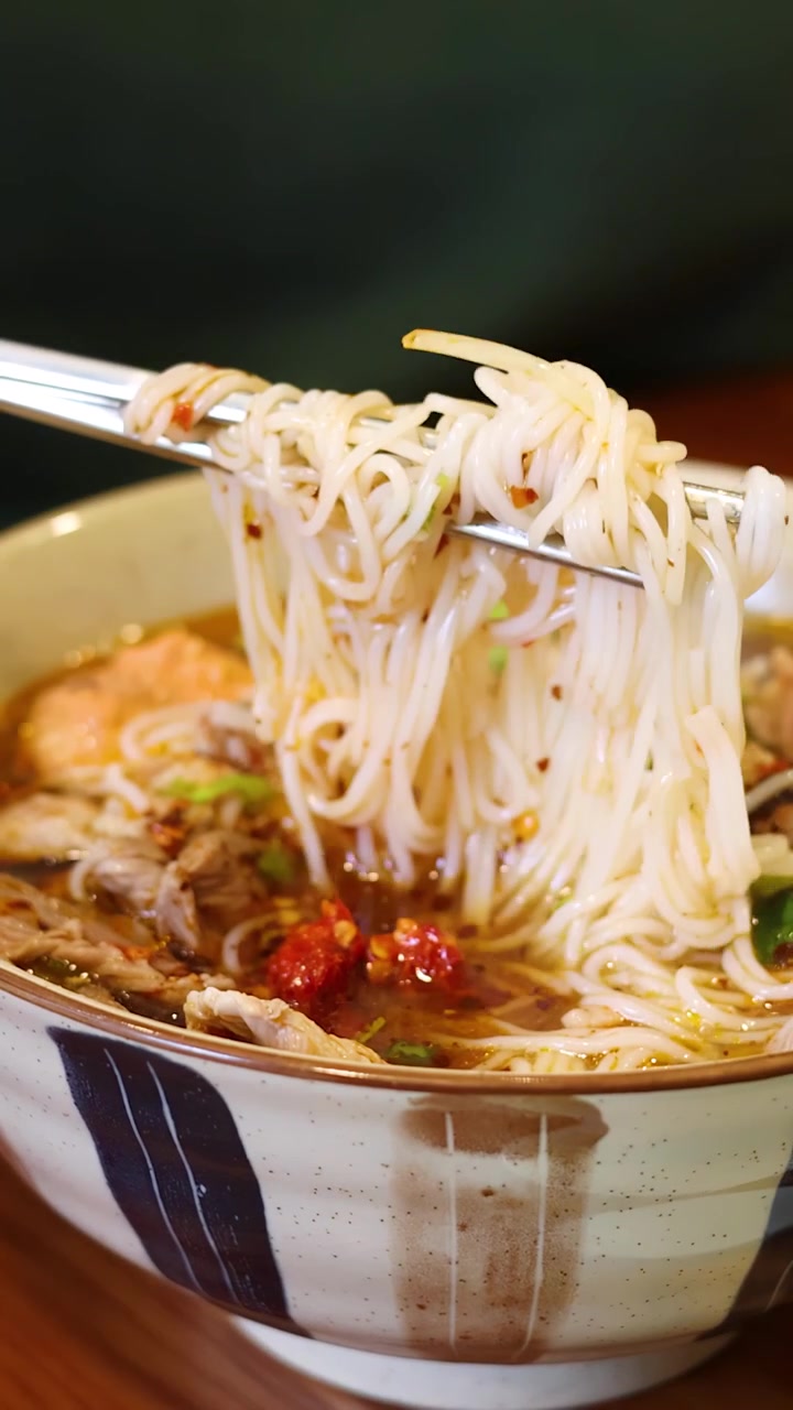 Una persona usa bacchette di metallo per sollevare spaghetti di riso da una ciotola di ceramica di zuppa.