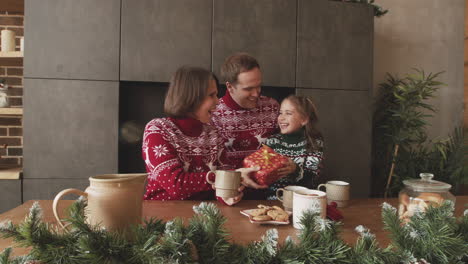 loving parents giving surprise gift to their little daughter while having breakfast together with hot drinks and cookies on christmas day at home