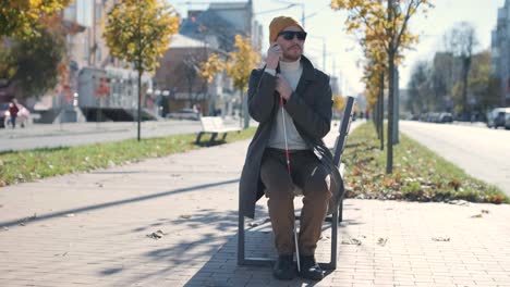 young blind man with smartphone sitting on bench in park in city, calling